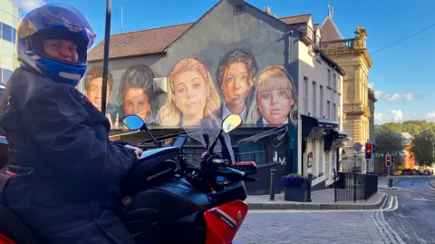 A turista Isabelle Lang fotografada em sua motocicleta, em frente ao mural Derry Girls.