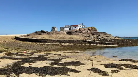 A coastal landscape showing a cluster of white buildings with red and grey roofs perched on a rocky outcrop surrounded by shallow water. In the foreground, a sandy beach is scattered with dark seaweed and tidal pools. A low stone walkway/seawall curves across the scene toward the rocky islet. The sky is a vivid, cloudless blue, and the bright sunlight highlights the textures of the rocks, sand, and buildings.