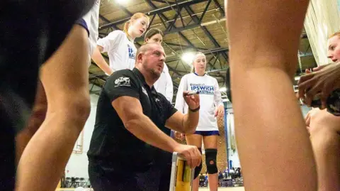 Robert Leach Head coach Nick Drane, crouching on the ground dressed in a black polo shirt and black bottoms delivers a team talk to surrounding players, who are mostly standing up