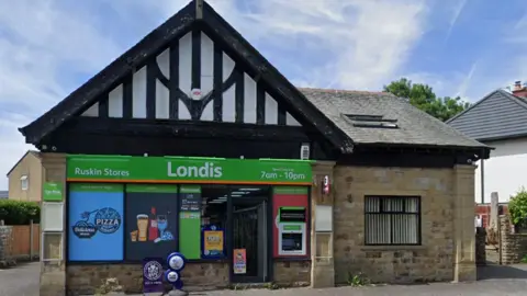 Street view image of the shop. A single storey stone building with a black and white Tudor-style pointed roof over windows covered in bright signage