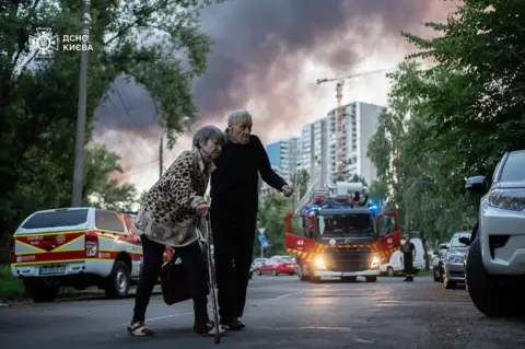 Woman and man with ambulances and smoke seen behind them