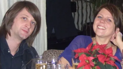 Zoe Alexander Nick and Zoe Alexander, sitting at a table, Nick is to the left with long dark hair, Zoe is to the right, with dark hair tied back. There are glasses and a plant in front of them. 