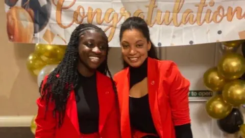 Elda Britto Two women are standing in front of a large "Congratulations" sign framed by gold balloons. The woman on the left has long black hair and is wearing a red jacket and a black T-shirt, while the woman on the right has black hair tied back and is wearing a red jacket with a black shirt. 