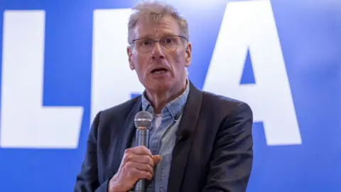 PA Media Kenny MacAskill, a grey-haired bespectacled man, speaks at an event while holding a silver microphone in front of his chest. He is wearing a dark jacket over a denim shirt. A blue Alba sign is in the background. 