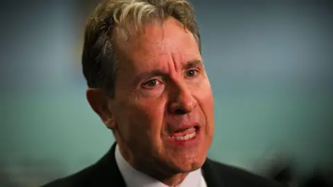A head and shoulders shot of Dan Norris wearing a white collared shirt beneath a black blazer. 