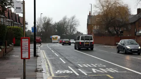 Bus and cycle lanes on Gosforth High Street, Newcastle. There is a shelterless bus stop at the left hand side, and on the road next to it is a white painted cycle lane, and then a white painted bus lane. There are a few cars driving at the right-hand side. Houses are also visible on either side of the road. It is a dull day with a grey sky, and there are some bare trees near the houses on both sides.