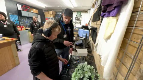 Antony Baker pictured in a black nike cap and black hoodie, teaching a young boy with blonde hair. The young boy has black headphones over his head and is wearing a black puffer jacket. They are both above DJ mixing decks