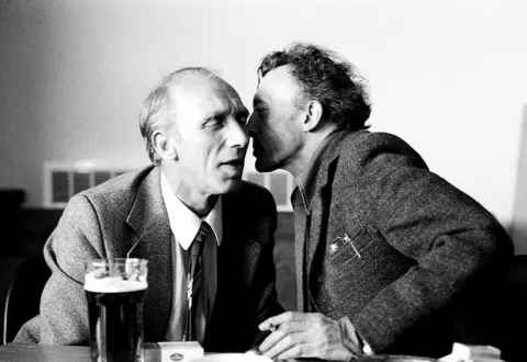 Ella Murtha A black-and-white photo of two men. The man on the right has brown, short hair and is leaning into the other man. It looks like he is either going to whisper something in his ear, or kiss him. The man on the left is fair and balding and has his eyes closed and mouth open. They are both wearing suits and there is a pint of stout on the table in front of them.