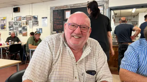 A man with a bald head, glasses and white checked shirt sitting in a cafe with other men

