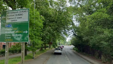 Google A road, with cars lining up at traffic lights, heading away from the camera. To the left are houses, trees and a large green road sign, while to the left there are trees and a thick green hedgerow