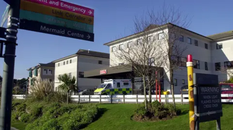 The photo shows the exterior of the Royal Cornwall Hospital in Truro, on a sunny day. A sign showing directions is visible in the foreground while an ambulance is seen parked in front of the building.