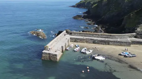 Gorran Haven harbour viewed from a drone on a sunny day. There are eight small motorboats tied up and there are two laid up on the pale sand. The harbour is two straight granite walls which meet at a 90 degree angle to protect the beach. Theer are large rocky outcrops in front of it and beyond. The rugged coastline has steep rocky cliffs which meet the water. The sea looks calm and dark seaweed can be seen as it is so clear. 