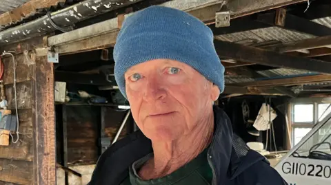 Tom Jackson/BBC Steve Wale is stood in the middle. He is wearing a blue had and a navy coat and is stood in a wooden building. 