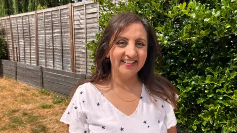 Bep Sandhu standing in a garden she is smiling at the camera. She wears a white top with small dark stars on and a thin gold necklace.