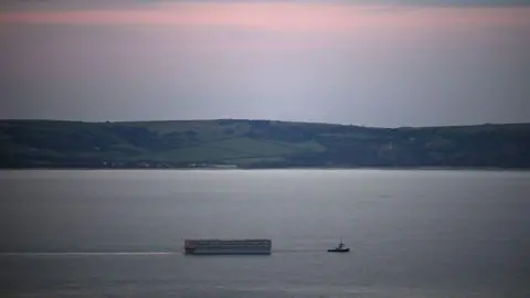 Finnbarr Webster/Getty Images The Bibby Stockholm is seen from a distance, being pulled through the sea by a tugboat. In the background are country hills and a pink sunset.