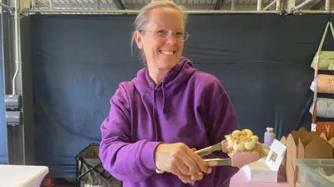 A woman wearing glasses and a purple top is smiling and looking to the left of the camera. She is holding a cake and a small white box is open on a table behind her.