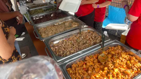 6 silver trays are filled with a variety of hot meals. Three people who are serving the food have red t-shirts on. Some customers can be seen queuing up to order.
