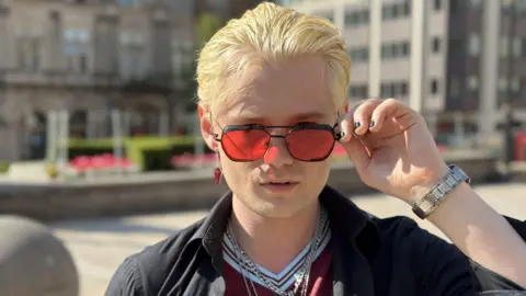 A close-up photo of a man with light blond hair, pink sunglasses, and an Aston Villa shirt.