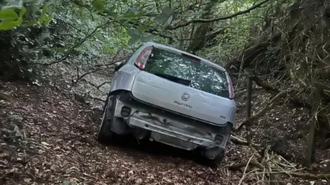 A silver Fiat Punto car left damaged and abandoned in a woodland area. The car is missing its rear bumper and number plate and is amongst trees and branches. 
