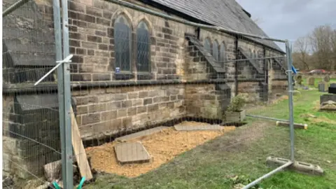 Barbara Gow A wall of the church with railings around an area which has two slabs ready for a memorial to be installed 