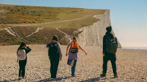 Rear view of a group of hikers walking across a beach toward white cliffs on a sunny day