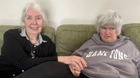 Judith and Monica are sitting on a green couch. Judith on the left has grey hair and is smiling at the camera, she has glasses on a chain round her neck. She is wearing a black cardigan with a black and white striped shirt underneath, she's holding her daughter's hand. She has grey hair and is wearing a dusty lilac hoodie with the word "Hamptons" written in white across it.