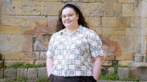 An individual stands in front of a textured stone wall, wearing glasses and a patterned short-sleeve shirt with heart motifs.

Curly hair is held back with a dark headband, and the person’s hands are placed casually in pockets.

The background features weathered bricks and small patches of greenery near the base.