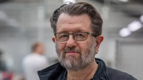 Northumbria University John O'Rourke is a man in his 50s with a grey beard and wearing glasses. He has brown hair and a blue top and the shot is of his head and shoulders 