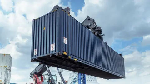 A dark blue shipping container being lifted in a dock in front of a cloudy sky