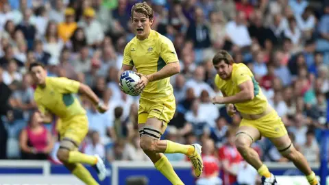 Getty Images An Australian rugby sevens player runs forward holding the ball, with two team mates behind him. They are wearing a strip that is nearly all yellow, with a little green on the jersey.