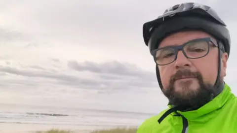 Family A man wearing a cycling helmet, glasses and a green coat, takes a selfie with the beach and sea visible in the background.