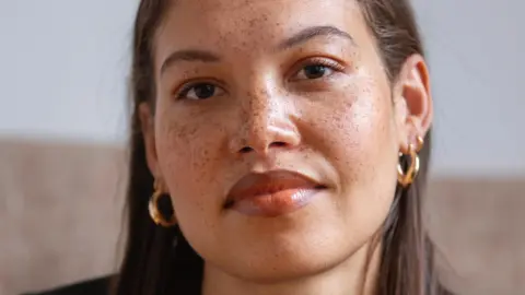 Seren Jones, who is of mixed race and has long brown hair, smiles at the camera. She is wearing gold hoop earrings and has freckles.