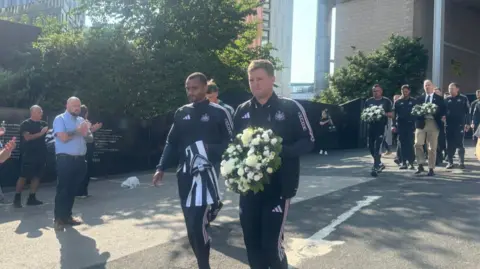 Newcastle United head coach Eddie Howe walks with club players and staff carrying a floral wreath of white and cream flowers with green foliage for Kevin Keegan at St James' Park. Several bystanders clap as he walks by.