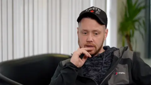 A man dressed in a grey tracksuit with a black cap on