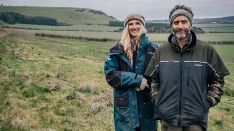 Greenshank Environmental A man and woman stand smiling on a field with a rural background and hills behind them. They are dressed in big jackets and it looks like a chilly day.