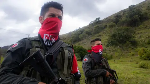 Getty Images Two fighters of the ELN with red and black face coverings and rifles in a forest clearing in the mountains of Colombia in 2025