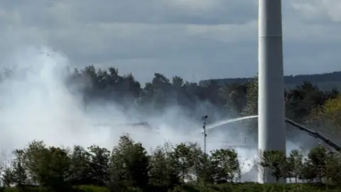 Smoke is rising from behind some trees with a fire engine spraying water on it.