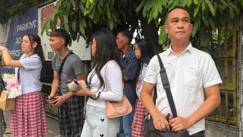 A man in a white shirt and a bag strapped across his shoulder stands on a road