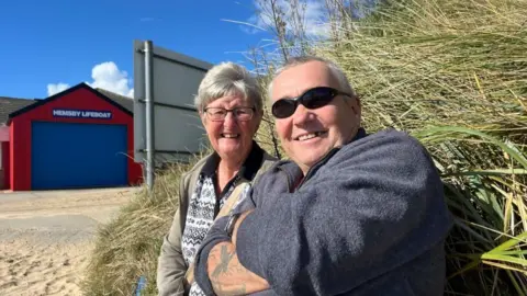 Andrew Turner/BBC John Peckham in a blue top with sunglasses and his wife Vanessa in a patterned top, sitting near the marram grass, with Hemsby Lifeboat Station in the background