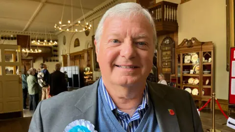 A man with short white hair smiles at the camera. He is wearing a blue and white striped shirt under a blue v-neck sweatshirt and a grey suit jacket. A poppy badge is on his left lapel and a blue and white rosette is on the other.