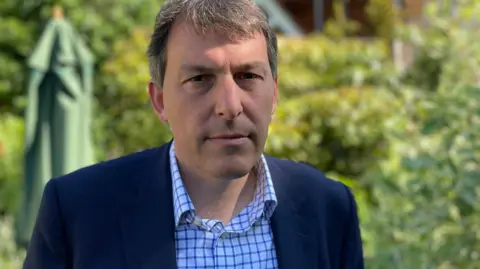 A man wearing a shirt and a jacket looks into the camera while standing in a garden.