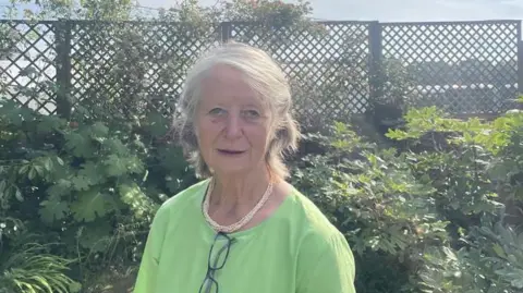 A woman wearing a green dress stands in a garden with bushes and a trellis seen behind her. She has a gold necklace on and her glasses hang on the top of her dress.