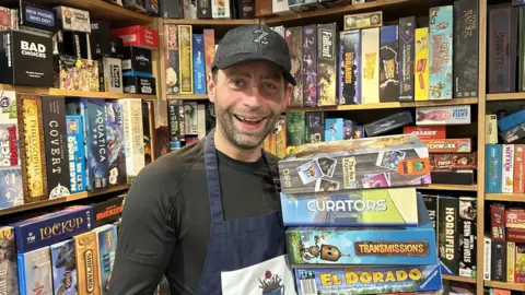 A man with a black cap, black top and navy blue apron holding a stack of colourful boxes in front of shelves filled with more colourful boxes