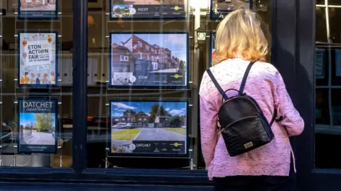 A woman wearing a pink jacket and a small black rucksack looks at adverts in an estate agent's window