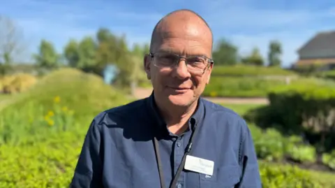 John Knight, who is wearing a blue shirt and a name badge. He is standing in a green garden.