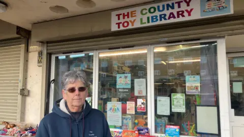 Nadia Lincoln/Local Democracy Reporting Service Heather Brakes with short grey hair and dark glasses wearing a blue fleece. She is standing outside the toy library that has various posters in the window and a table outside with books and toys.