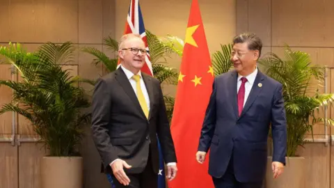 Prime Minister Anthony Albanese, wearing a black suit and a yellow tie smiled with the President of China, Xi Jinping, who is wearing a navy blue suit with a red tie. Both men are standing in front of their respective national flags. 