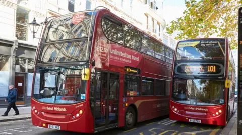 Two red West Midlands buses side by side on a busy city centre street. Both have West Midlands written in white on the front and adverts on the side. One has the number 101 on it sign as it stands at a bus stop. The other bus, to its right, appears to be driving off.