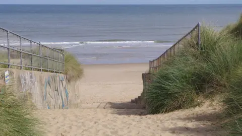 A route to a sandy beach can be seen, guided by two concrete walls of sea defences, looking from the dunes towards the sea on a sunny day