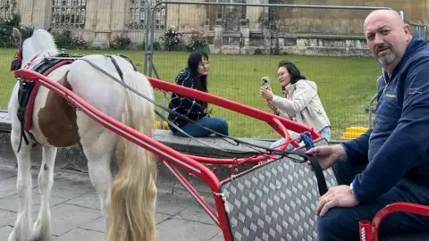 John Ball John Ball on a cart being pulled by a white and brown horse called Syble stop outside King's College in Cambridge. Two women sit on a wall nearby.